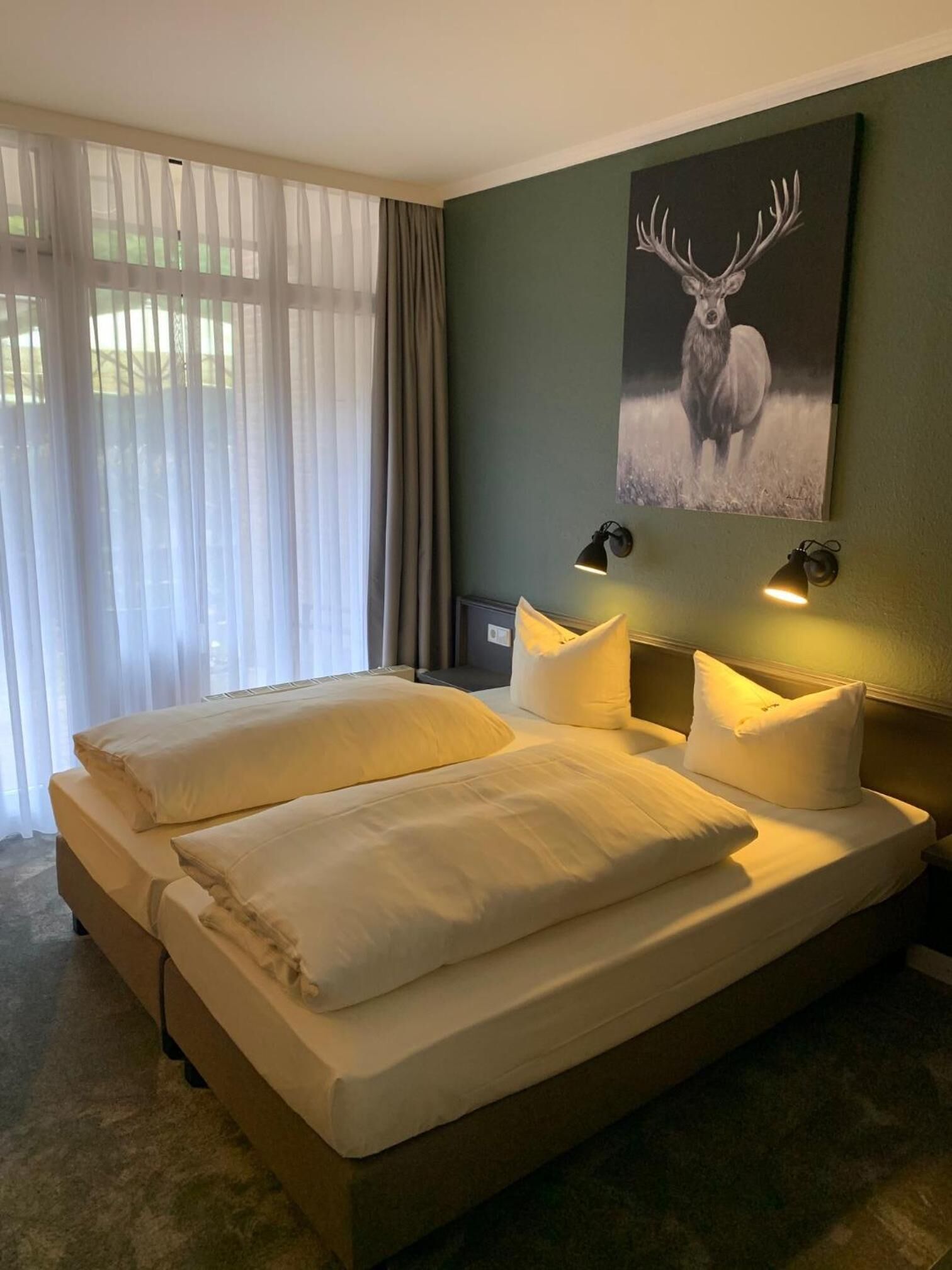 A bedroom with a large bed, two bedside lamps, a painting of a deer on the wall, and sheer curtains on the window.