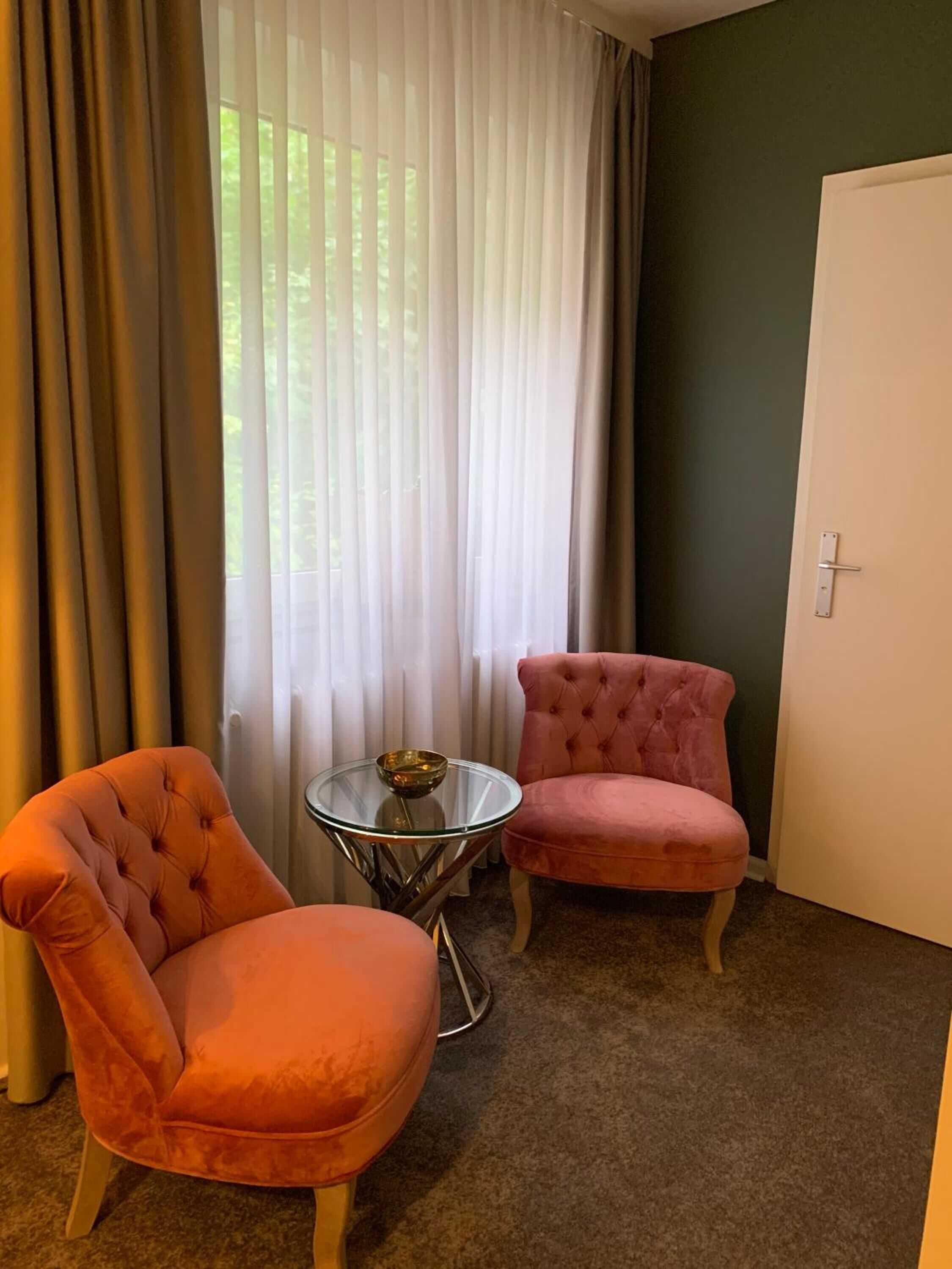 Une pièce avec deux fauteuils roses à motifs capitonnés, une petite table en verre sur laquelle se trouve un bol, et une porte blanche.