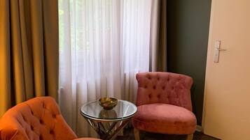 Une pièce avec deux fauteuils roses à motifs capitonnés, une petite table en verre sur laquelle se trouve un bol, et une porte blanche.