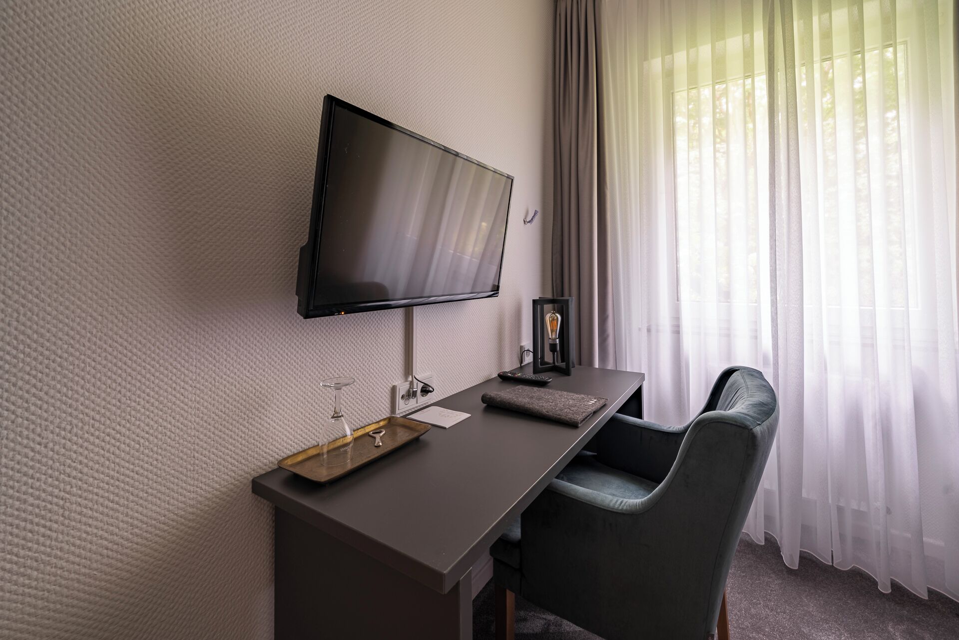 A modern hotel room with a flat-screen TV mounted on the wall, a desk with a remote control, a glass with a wine glass, and a chair.
