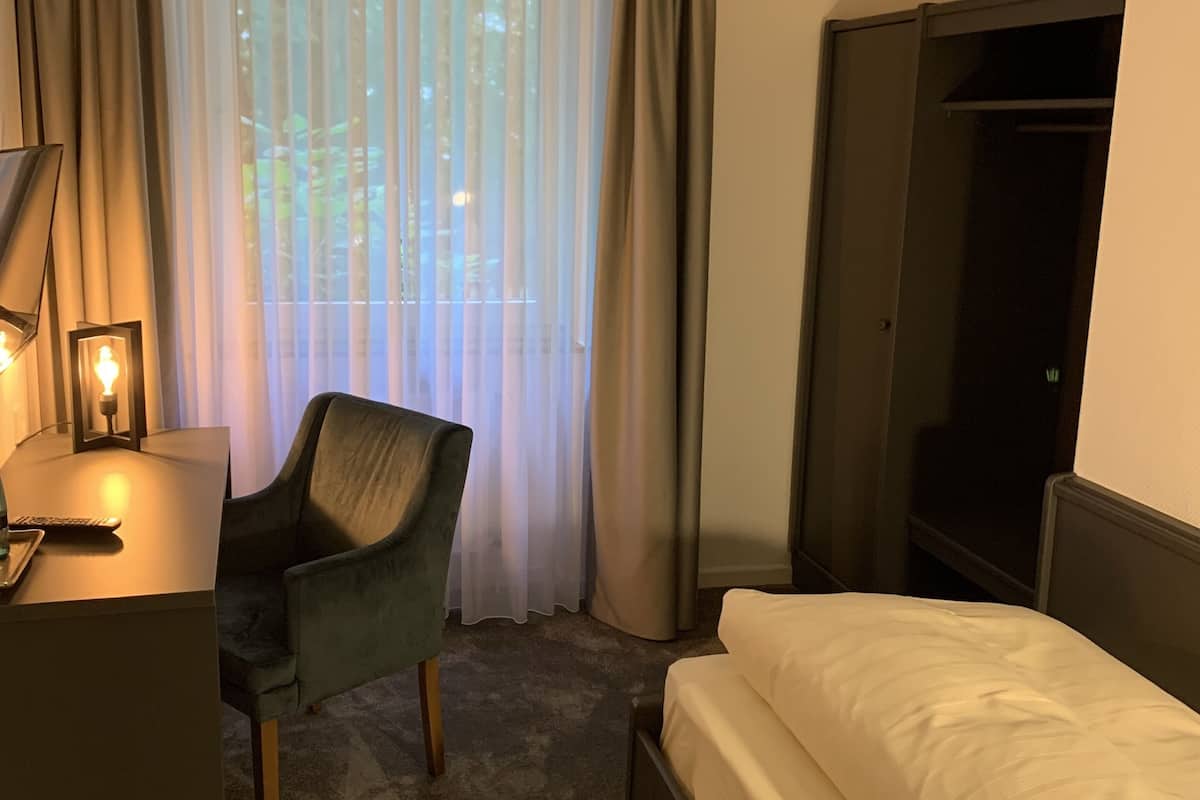 Quarto de hotel com uma cama, uma escrivaninha com abajur, uma cadeira e uma televisão.