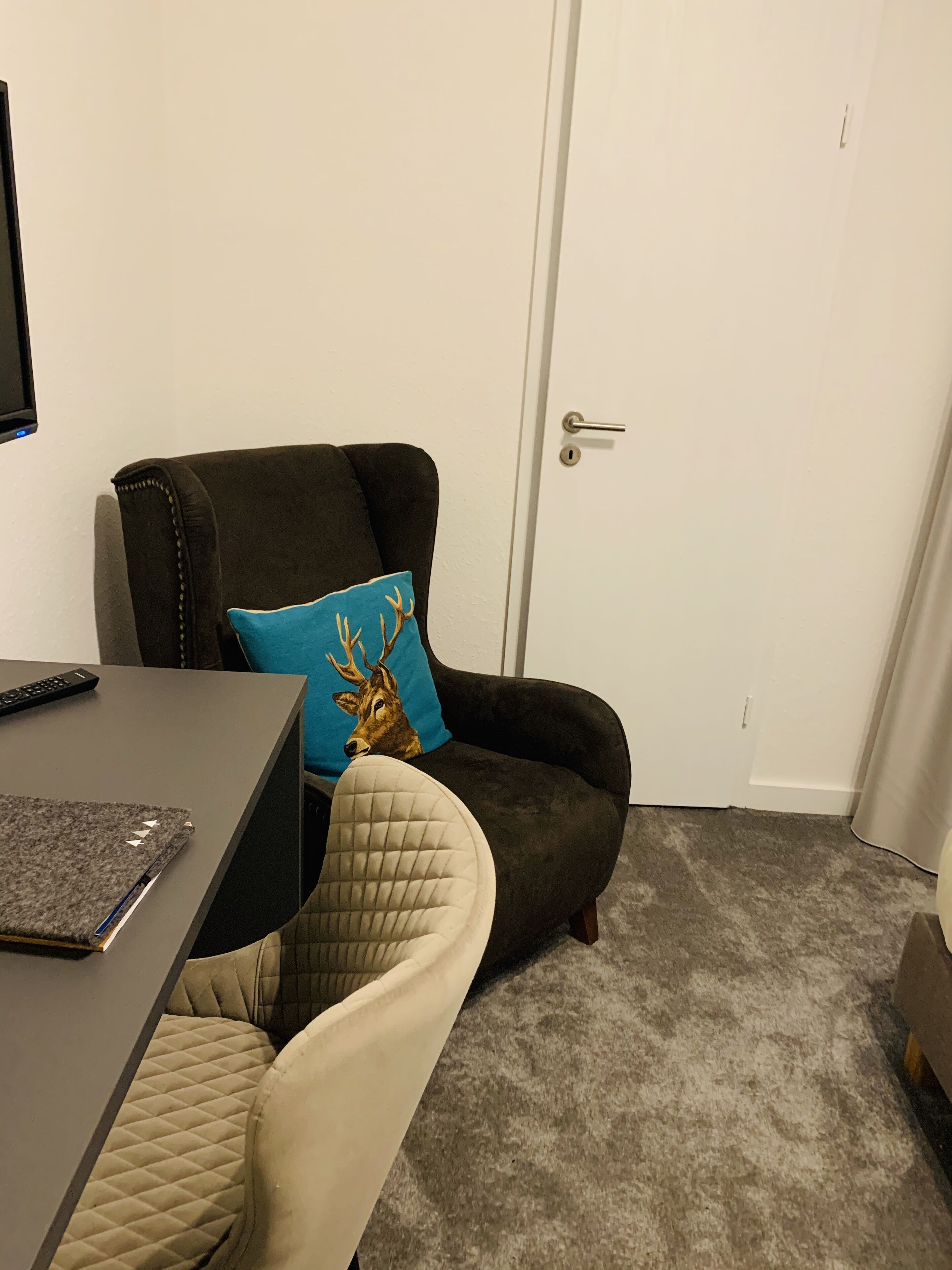 Una habitación con un sillón marrón oscuro, un sillón color beige, una mesita con un cuaderno y un control remoto, y una puerta con manija plateada.