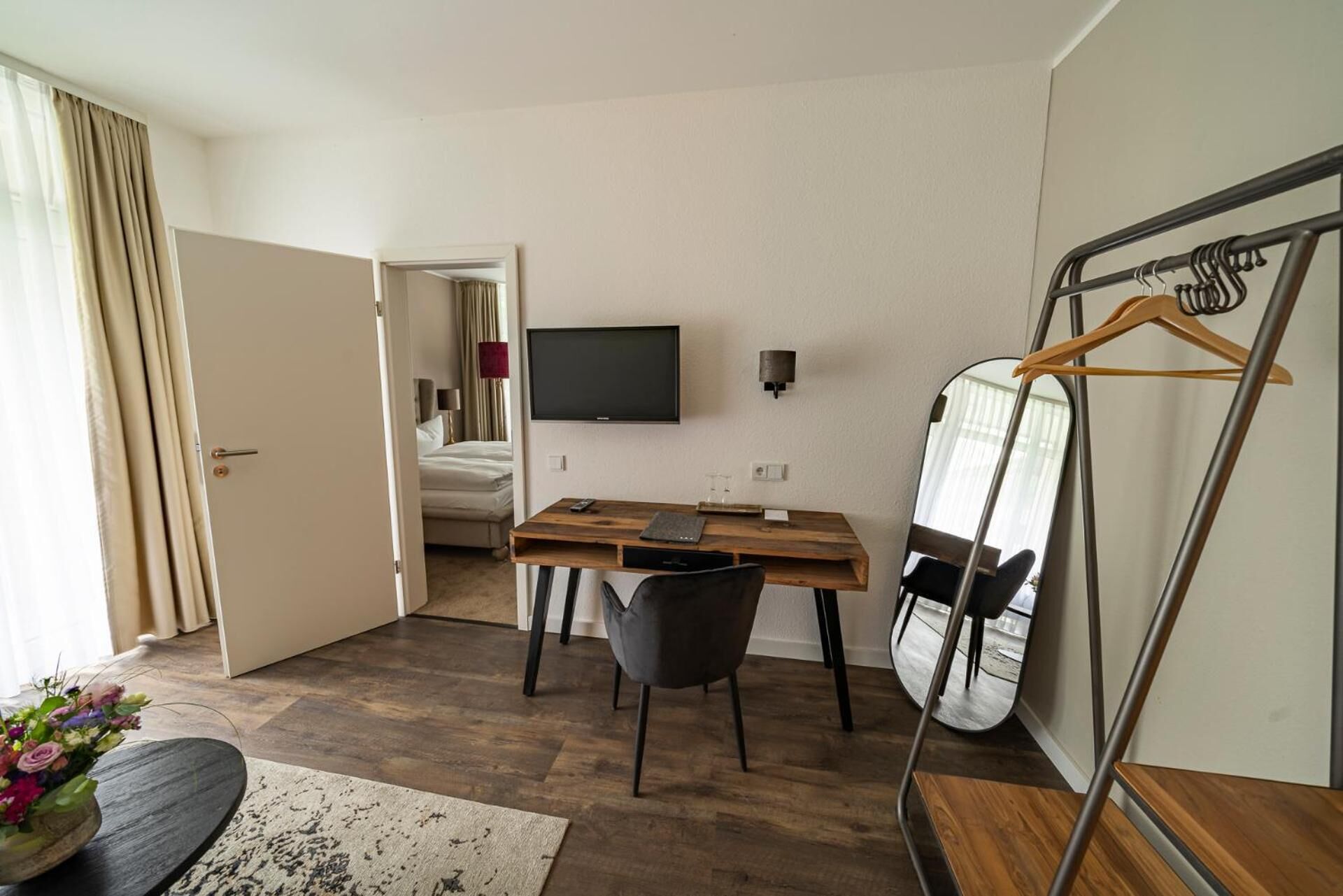 Ein Hotelzimmer mit Holzschreibtisch, einem Sessel, einem Bett, einem Fernseher, einem Spiegel und einem Kleiderhaken.