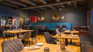 A restaurant with a modern interior, featuring wooden tables and cushioned seating. There are decorative wall sconces and a blue cushioned bench in the background. - Waldhotel Riesebusch (Bad Schwartau)