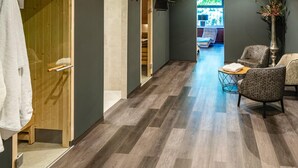 A hallway with wooden flooring, a seating area with patterned chairs, and a view into a room with a bed and a window. - Waldhotel Riesebusch (Bad Schwartau)