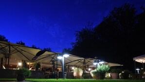 An outdoor dining area with multiple tables and umbrellas, surrounded by greenery and a building in the background. - Waldhotel Riesebusch (Bad Schwartau)