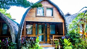 A wooden house with a thatched roof, a porch with a table and chairs, and lush greenery around. - Cotton Tree Cottages (Gili Trawangan)