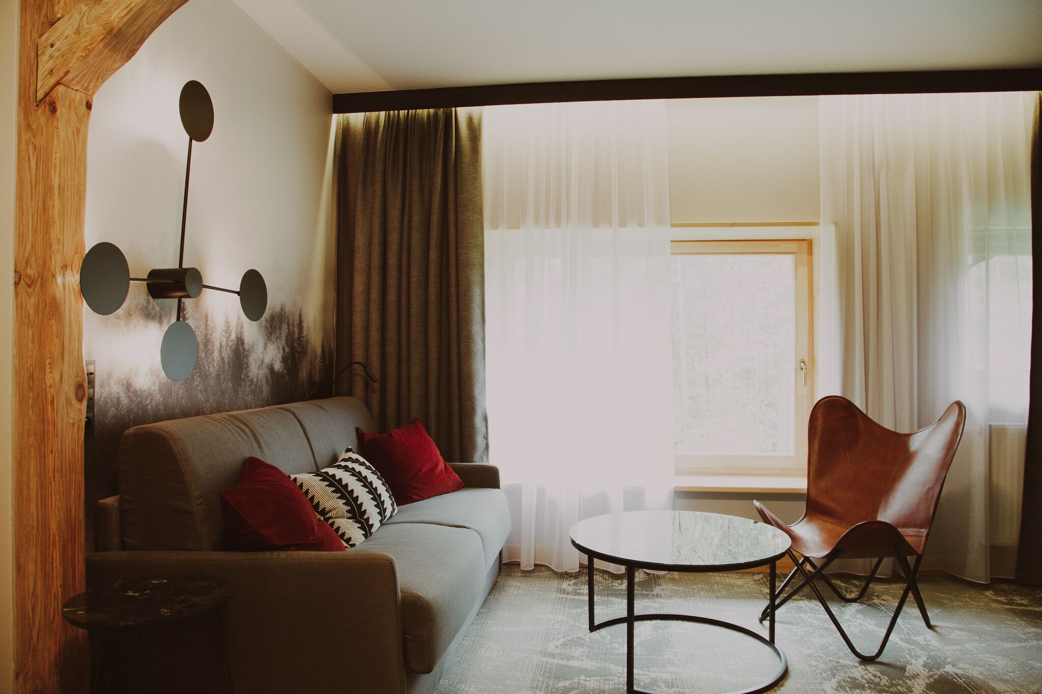 A living room with a sofa, a coffee table, and a chair. There is a window with curtains, a wall-mounted light fixture, and a radiator.