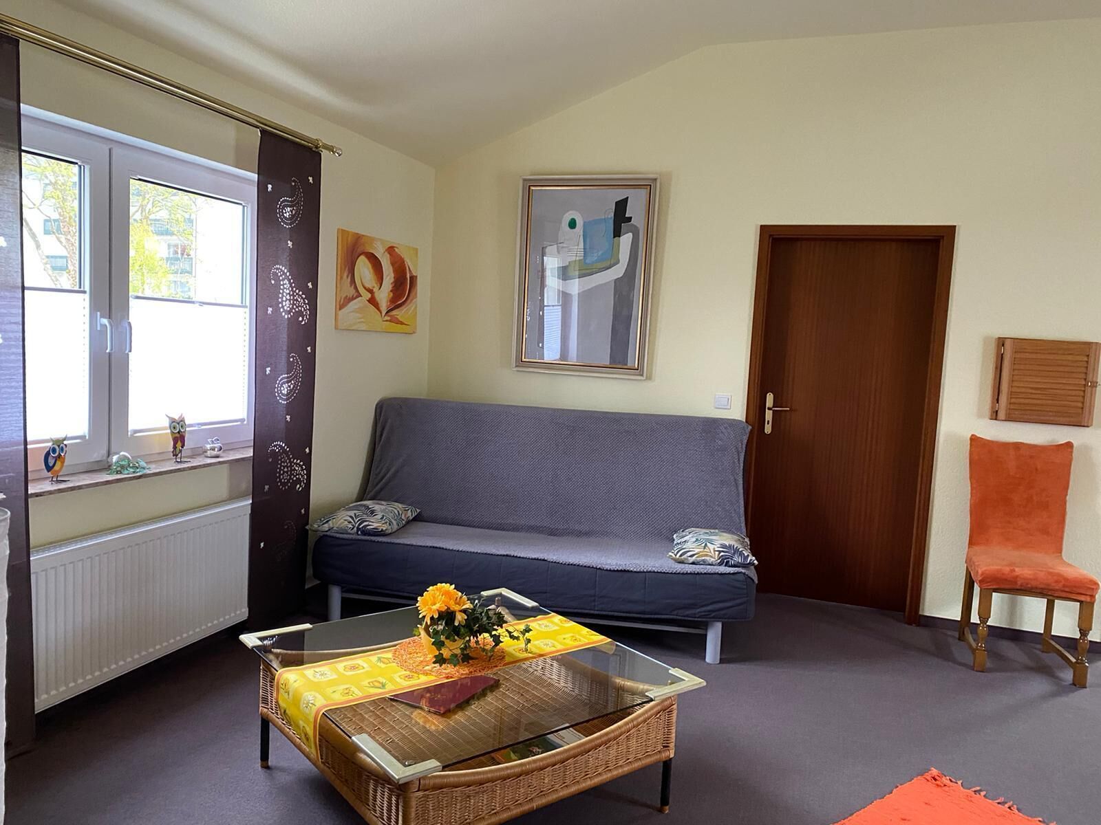 En stue med sofa, sofabord med glasplade og gul gennemsigtig dug, dør og vindue med gardiner.