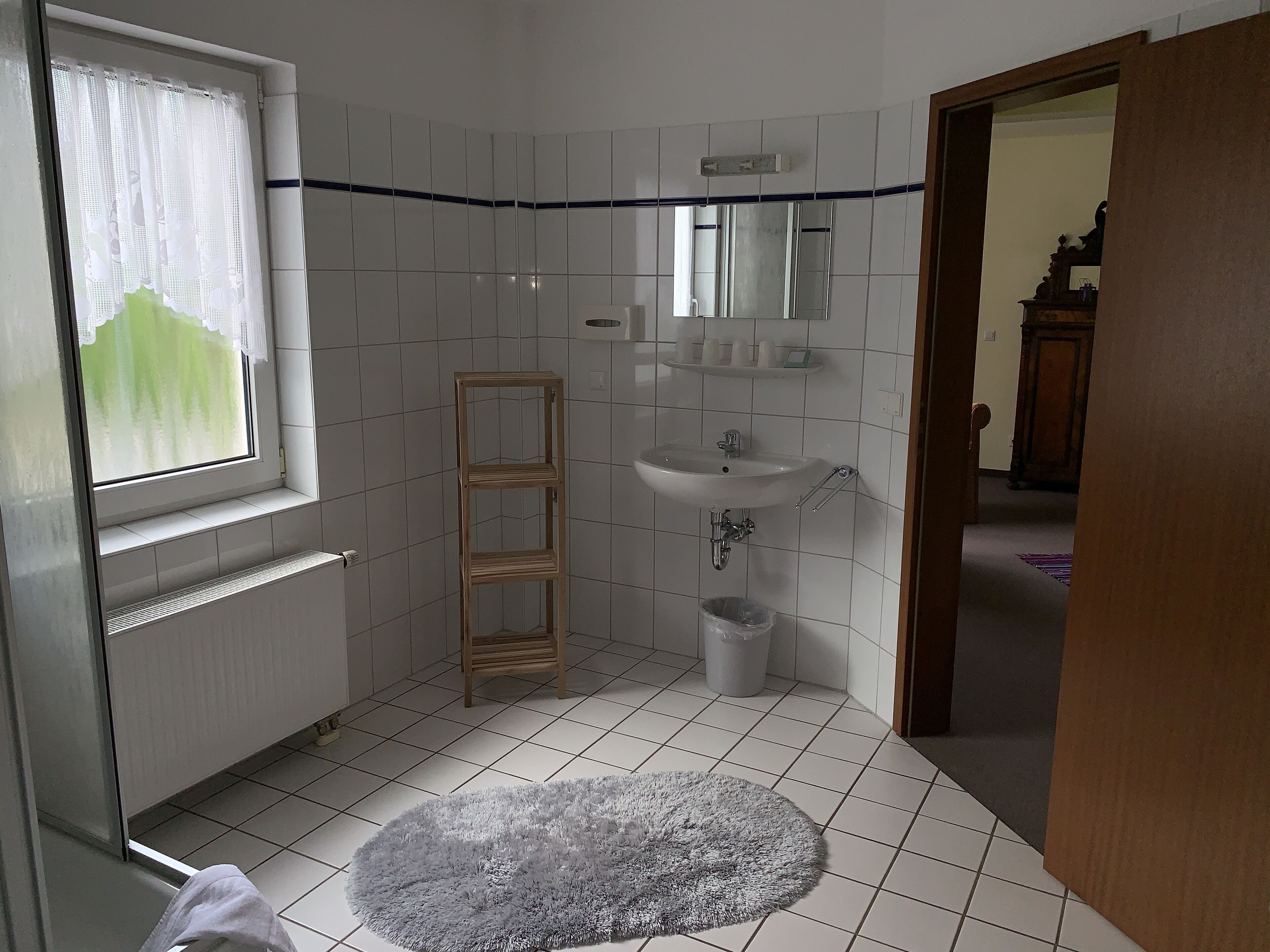 Une salle de bain avec une fenêtre, un lavabo, un miroir, une étagère et un tapis.