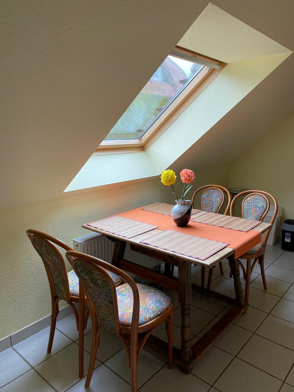 Un comedor con mesa y sillas, un tragaluz y un jarrón con flores.