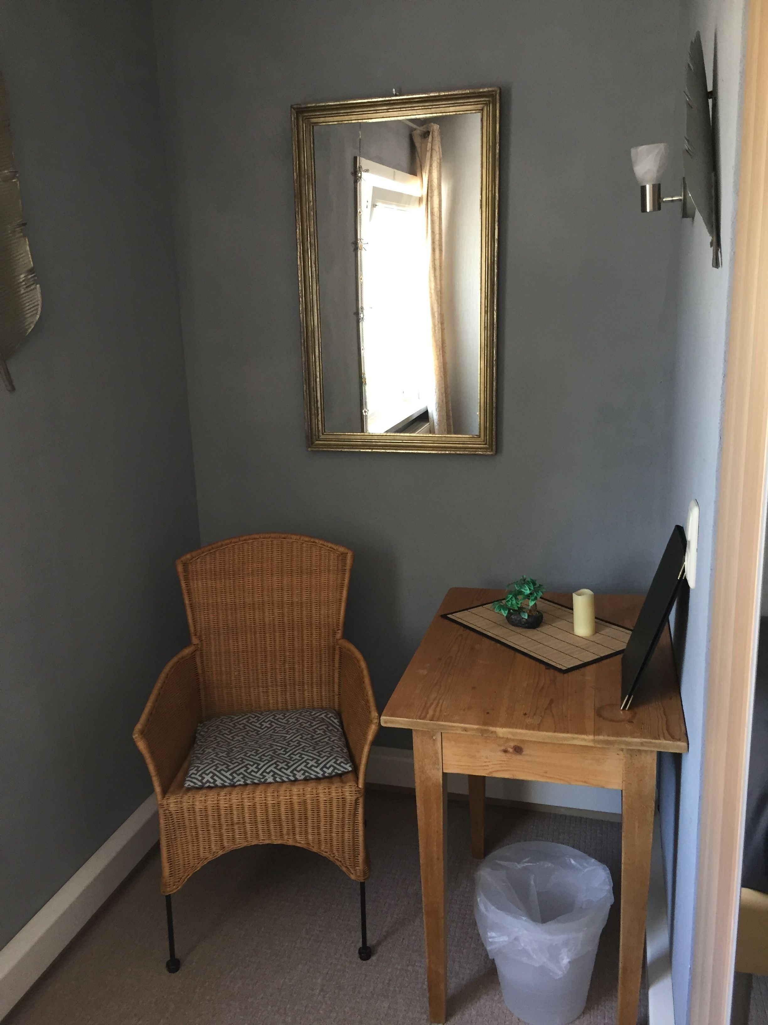 Una habitación con una silla de mimbre, una pequeña mesa de madera con una vela y una planta, un espejo en la pared y un cesto de basura.