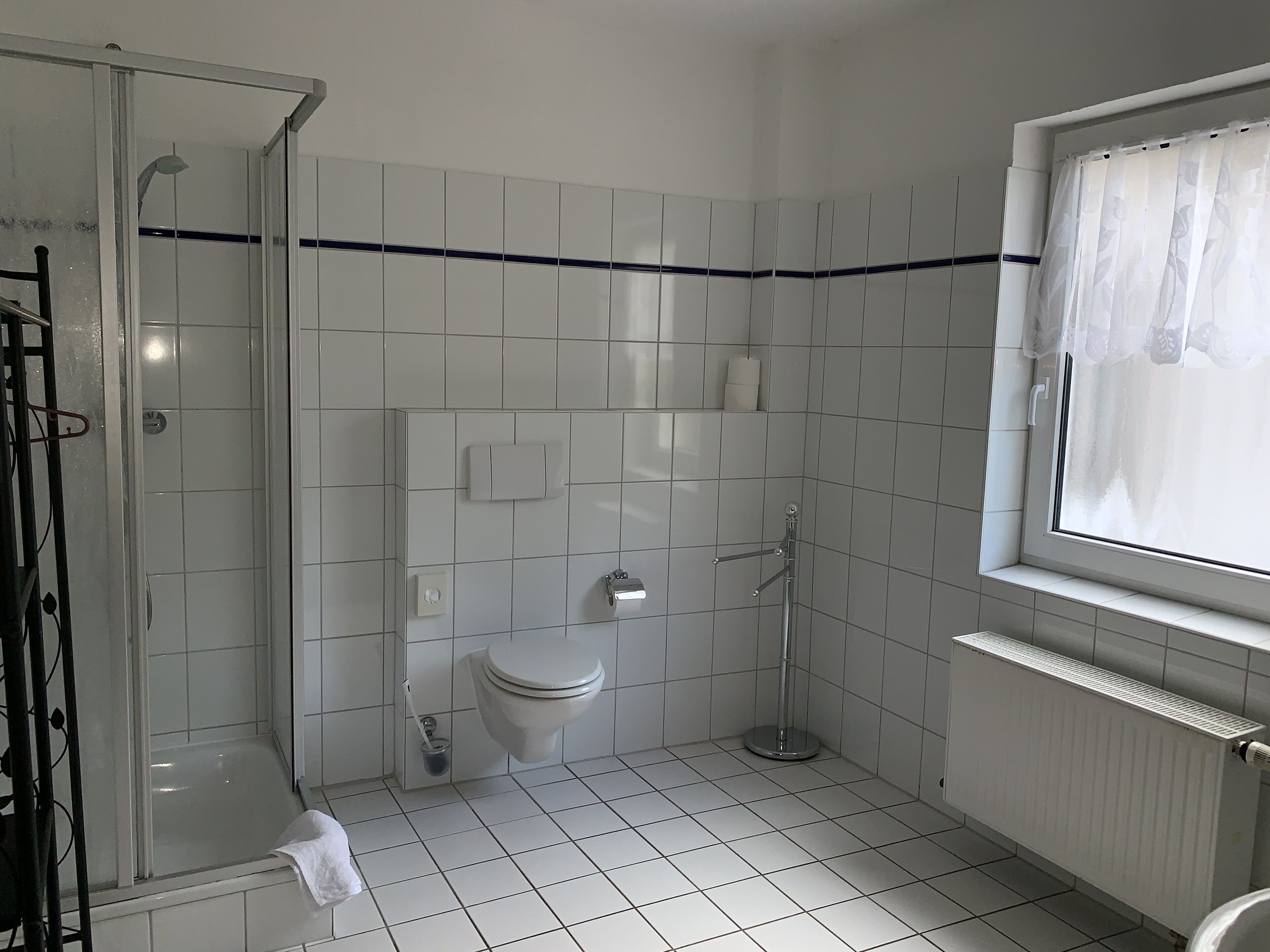 Bagno con piastrelle bianche, un water, una doccia e una finestra con tenda.