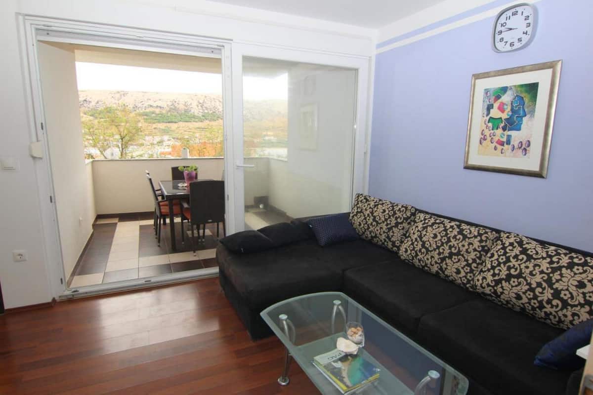 A living room with a black sofa, a glass coffee table, and a view through a sliding glass door to an outdoor balcony.