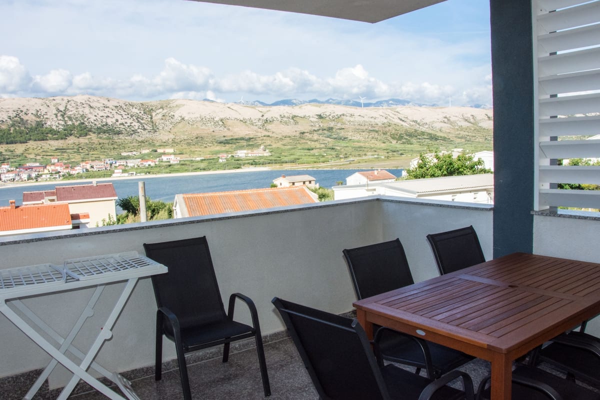 A balcony with a table and chairs, overlooking a body of water and a hilly landscape.