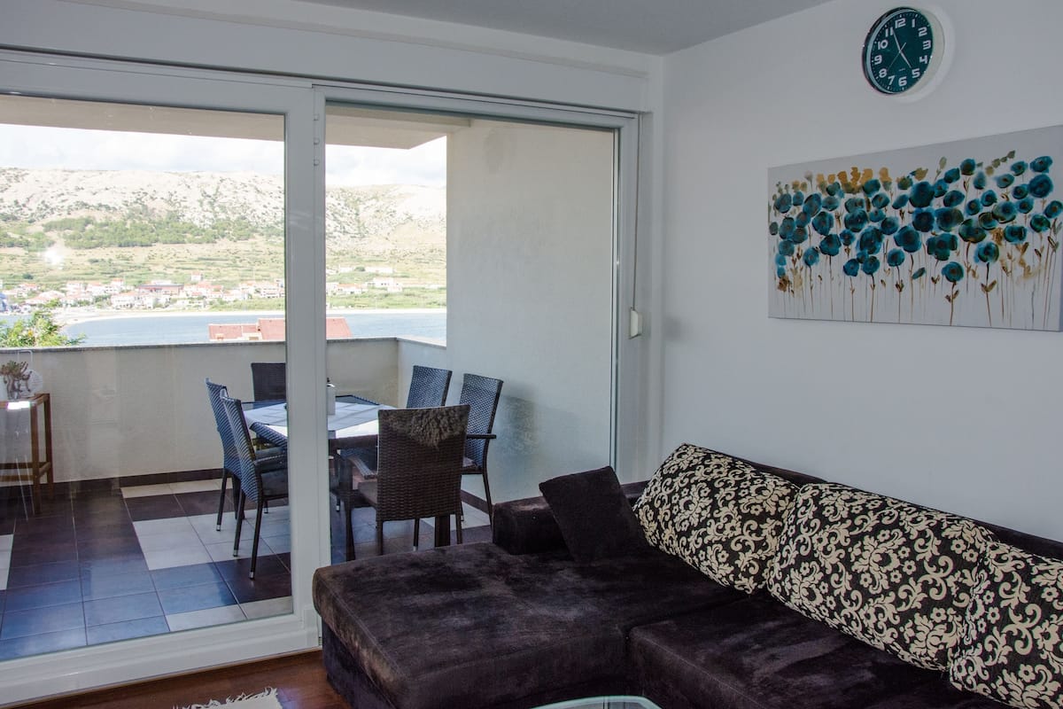 A living room with a sofa, a dining area with chairs and a table, and a view of a hillside through a glass door.