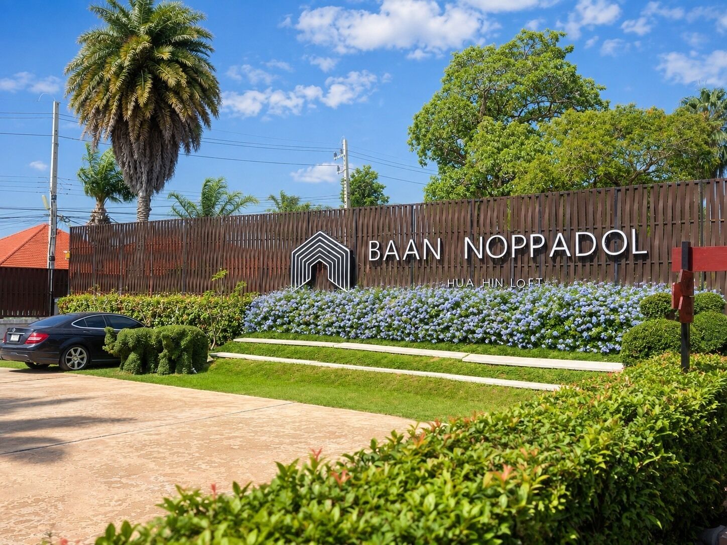 A residential area with a wooden fence, a car, and a sign that reads 'BAAN NOPPADOL.'