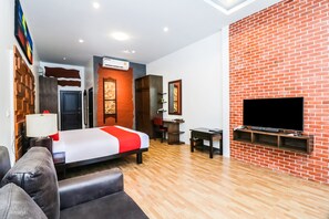 A modern hotel room with a brick accent wall, a flat-screen TV mounted on the wall, a bed with a red and white bedspread, a wooden floor, and a seating area with a sofa and armchairs. - Baan Noppadol Hua Hin Loft Hotel (Hua Hin)
