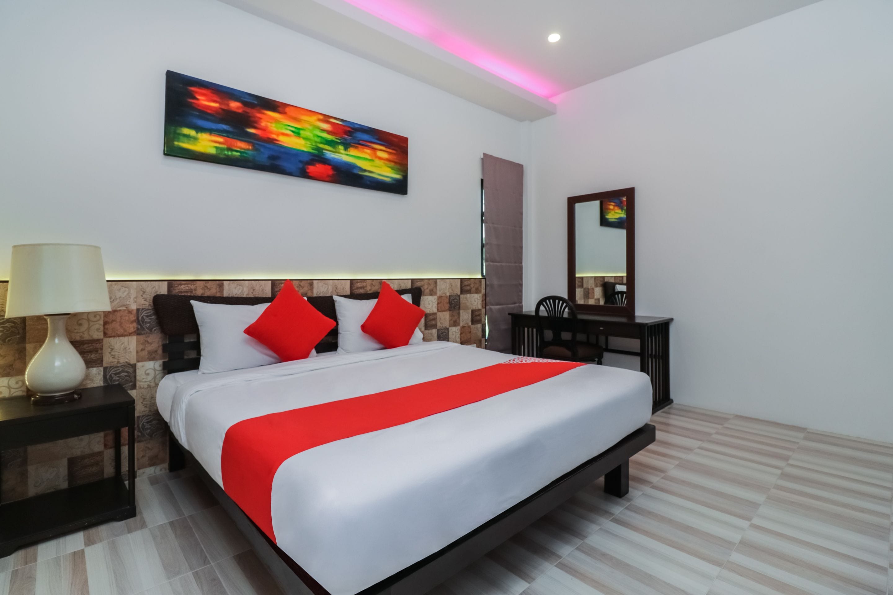 A modern hotel room with a large bed, a nightstand, a desk, a chair, a mirror, and a colorful abstract painting on the wall.