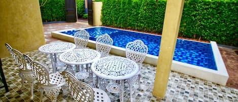 A pool area with white metal chairs and tables, surrounded by a tiled floor and a low hedge.