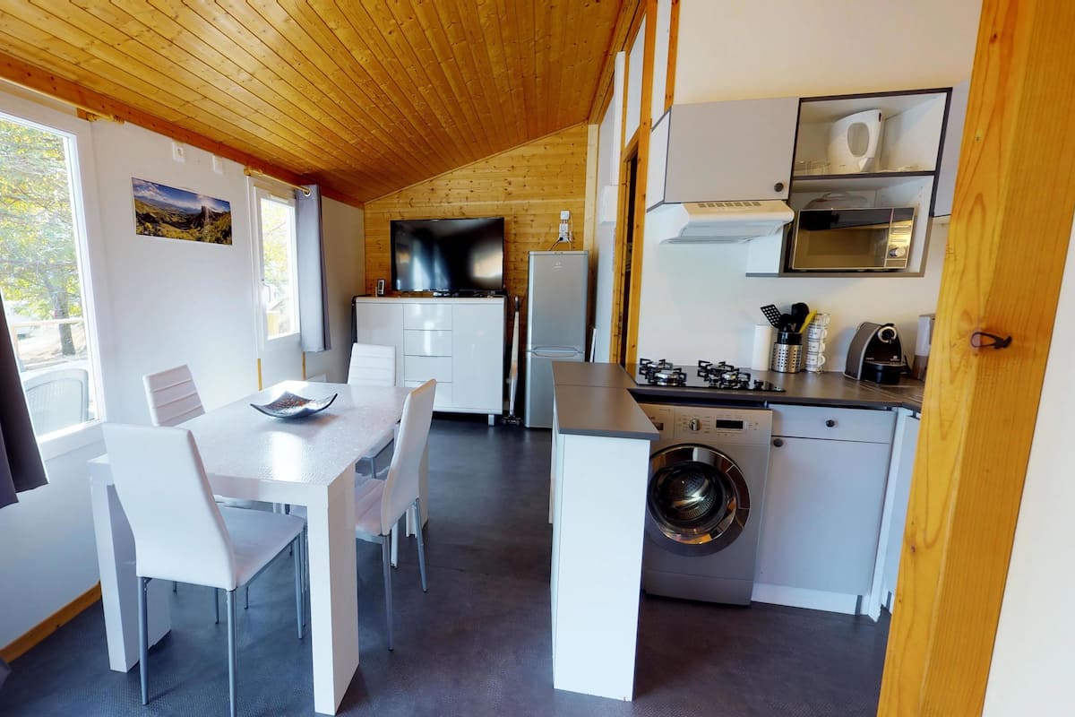 Cocina compacta con lavadora, zona de comedor con mesa y sillas, y un ventanal grande con cortinas.