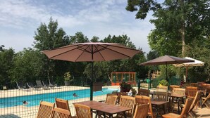 Un espace aménagé au bord de la piscine, avec des chaises et des tables en bois, des parasols et une piscine entourée d’une clôture.