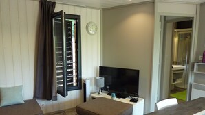 Un salon comprenant un téléviseur, un canapé, un pouf, une fenêtre avec des stores et une horloge au mur.