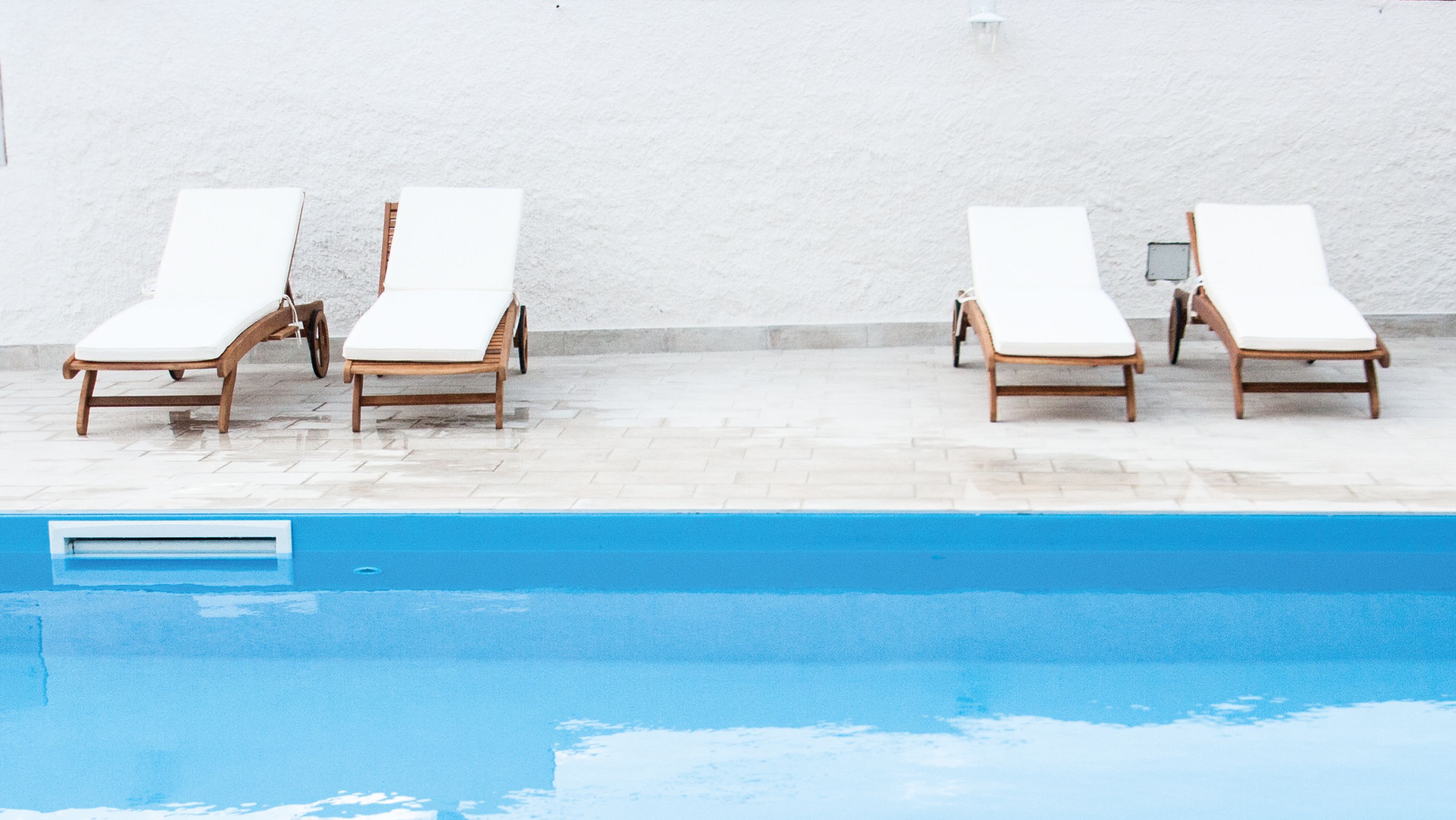 A swimming pool with four wooden lounge chairs aligned against a white wall.