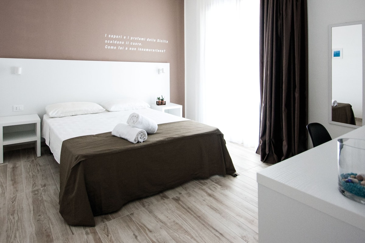 A modern hotel room with a large bed, a bedside table, a small table with a plant, and a mirror on the wall.