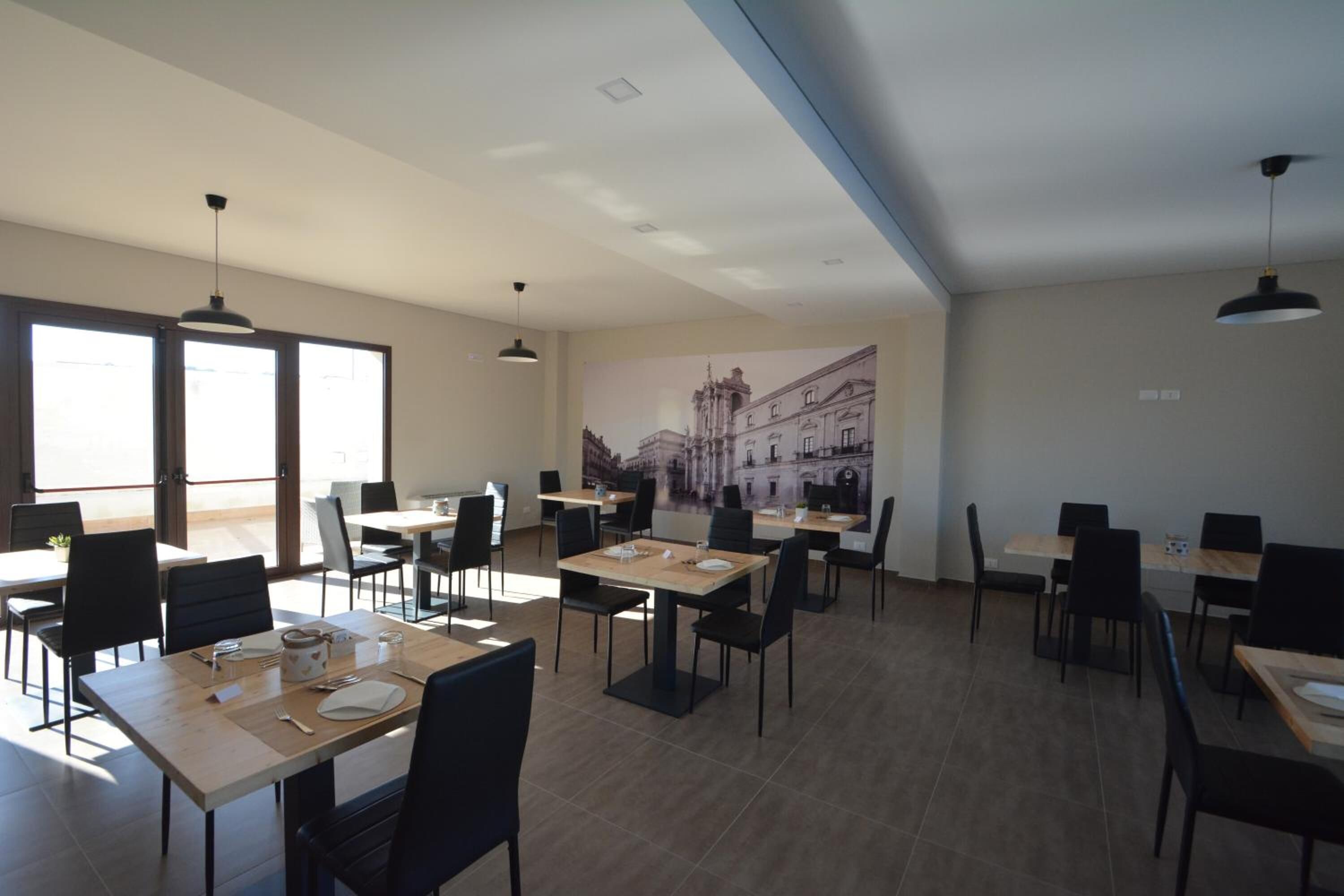 A spacious dining area with multiple tables and chairs, large windows allowing natural light, and a cityscape mural on the wall.