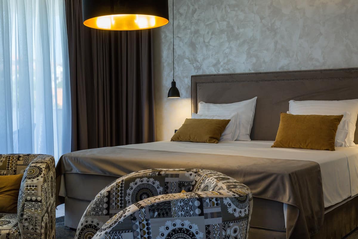 A hotel room with a large bed, a patterned armchair, a hanging lamp, and a view through sheer curtains.