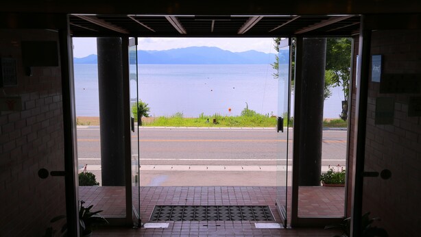 A view through an open doorway to a paved area with a metal grate, potted plants, and a distant body of water with mountains in the background. - Lake Side Hotel MINATOYA (Inawashiro)