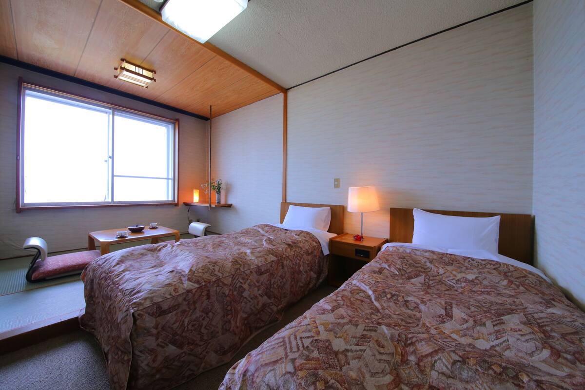 A room with two beds, a window, a bedside table, and a lamp.