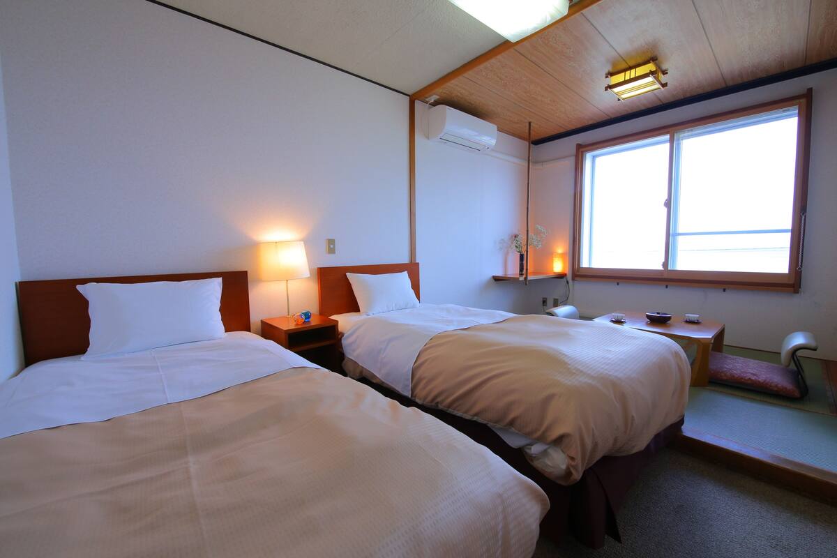 A hotel room with two beds, a nightstand, a small table, a chair, and a window with natural light.