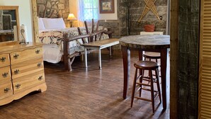 Une pièce rustique avec un mur en pierre, des meubles en bois, un lit recouvert d’une couverture à motifs, un banc, une table avec des tabourets et un tableau encadré accroché au mur.