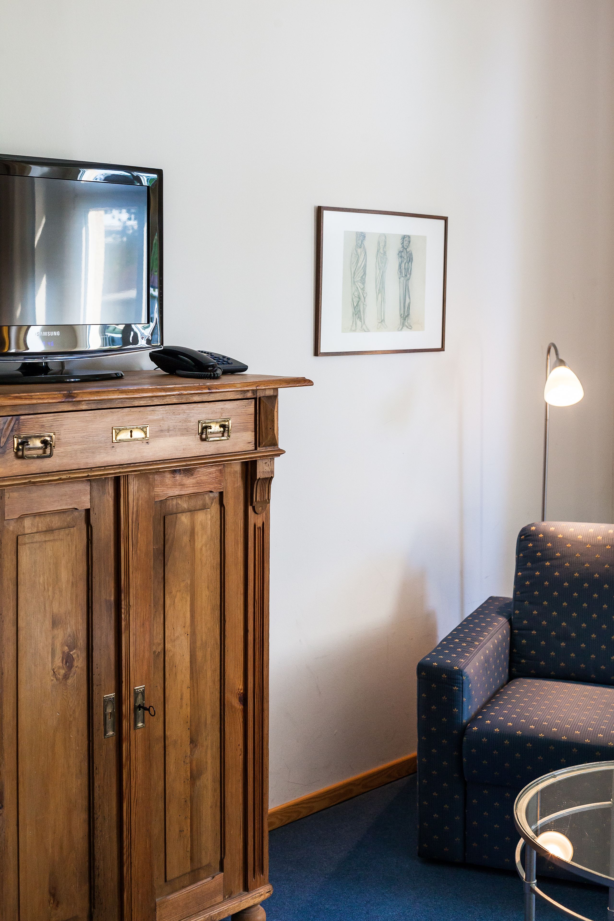 Un mueble de madera con televisor y teléfono, una obra de arte enmarcada en la pared, una lámpara y un sillón estampado.