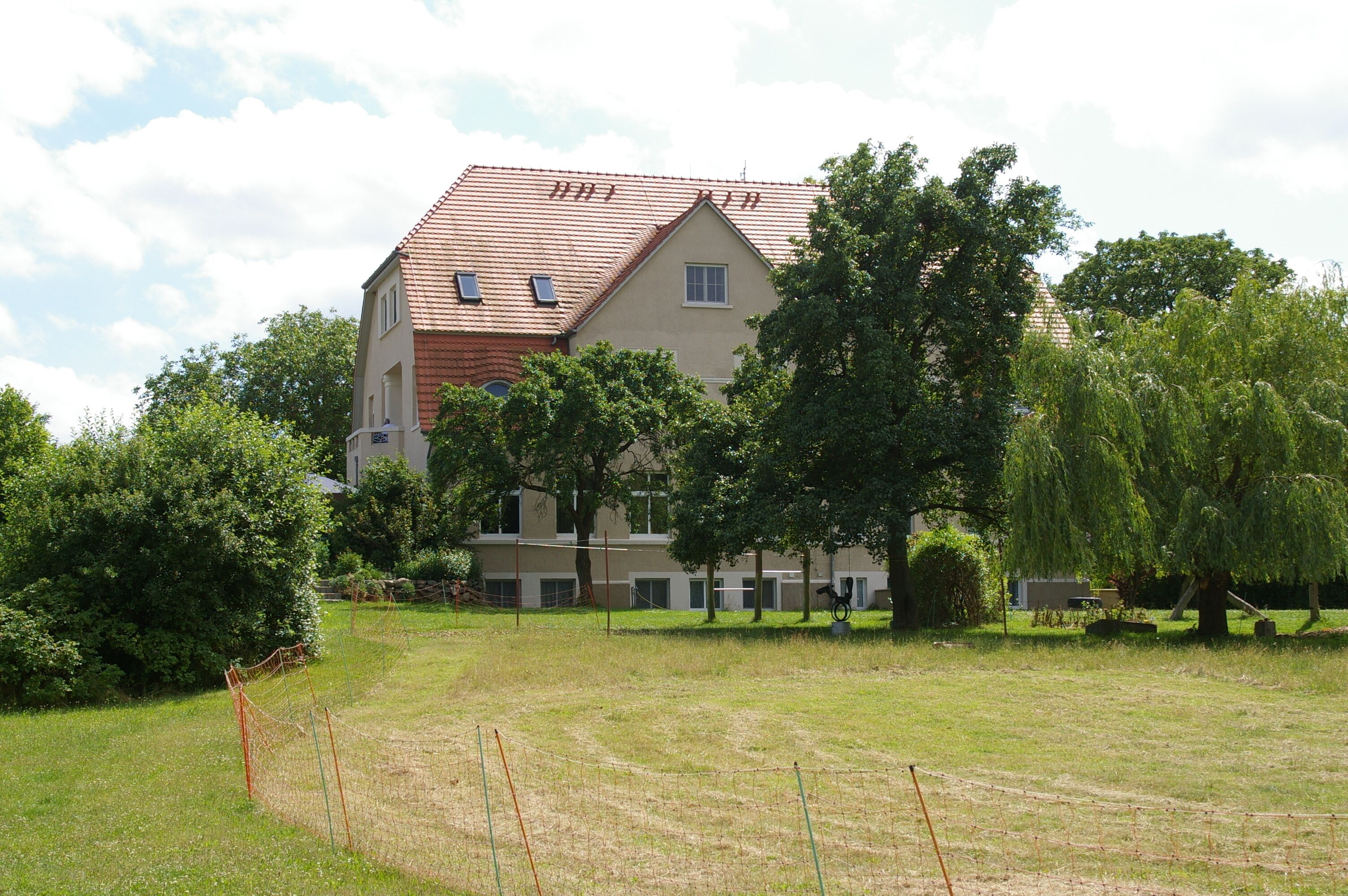 Un bâtiment à deux étages, avec un toit recouvert de tuiles rouges, entouré d’arbres et d’une zone délimitée par une clôture.