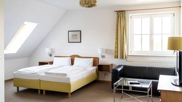 A hotel room with a bed, a black leather sofa, a glass coffee table, a desk with a lamp, a window with curtains, a framed picture on the wall, and a ceiling light fixture.