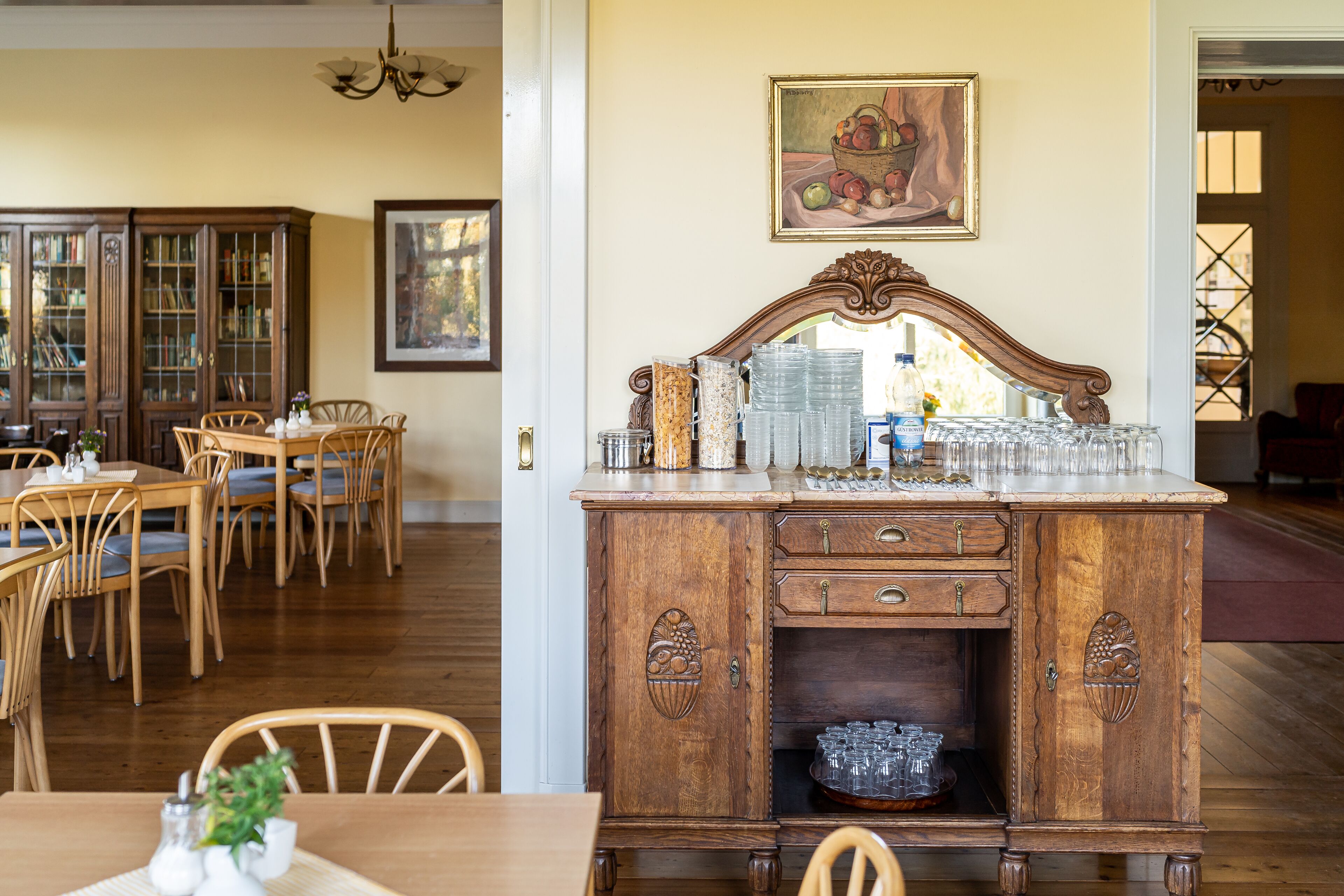 A dining area with wooden chairs and a table, a wooden cabinet with glassware, and a painting on the wall.