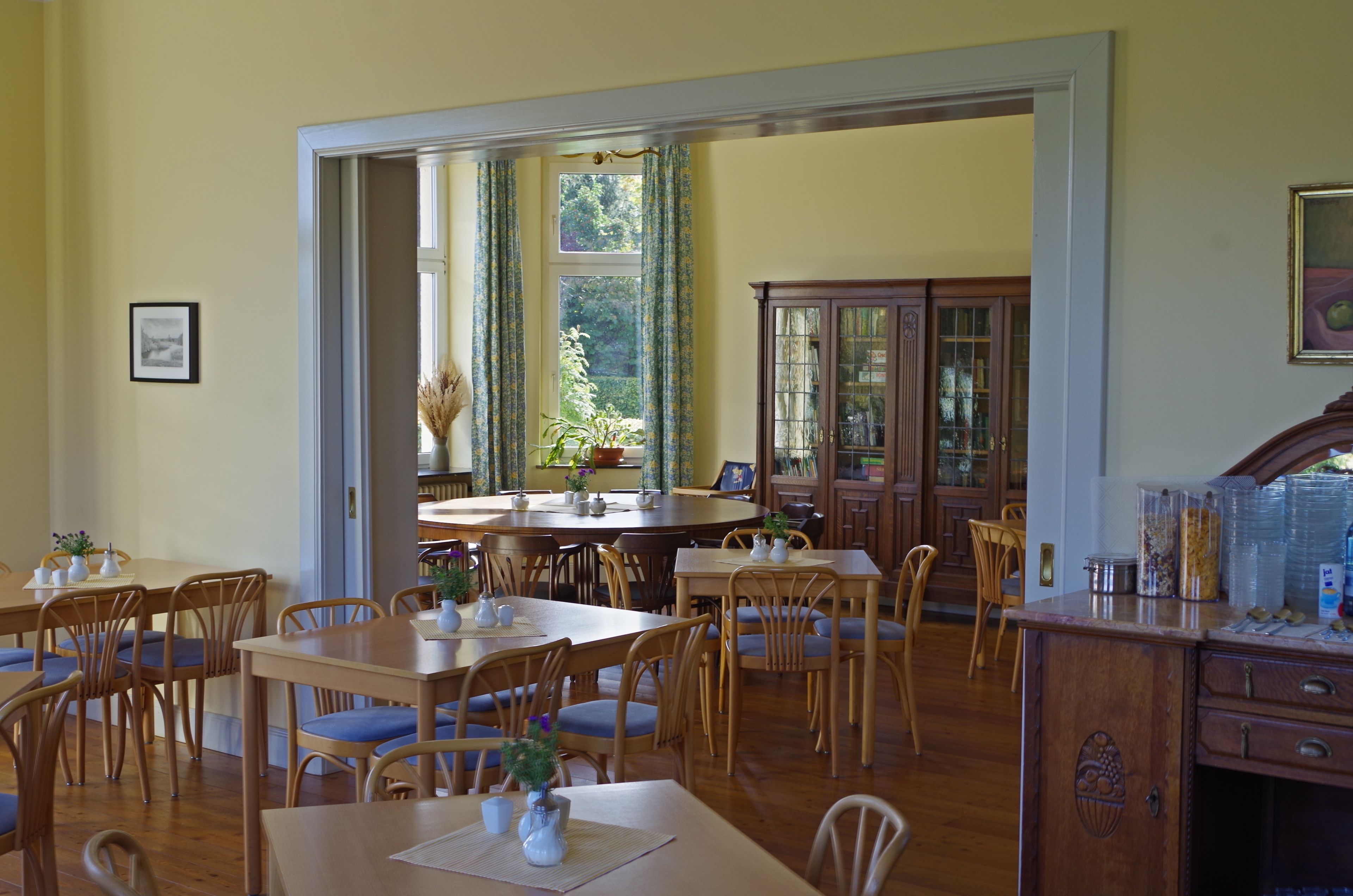 Un comedor con mesas y sillas de madera, un ventanal con cortinas y un aparador con puertas de cristal.