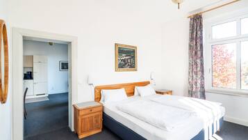 A bedroom with a large bed, wooden headboard, two bedside tables, a nightstand, a painting on the wall, and a window with curtains.