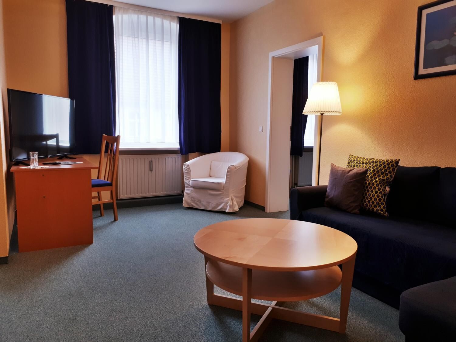 A hotel room with a TV, a round table, a sofa, a chair, and a lamp.