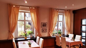A dining area with two tables, each with four chairs. There are potted plants on the windowsills, and the room has wooden flooring. - Hotel Krone (Lindow)