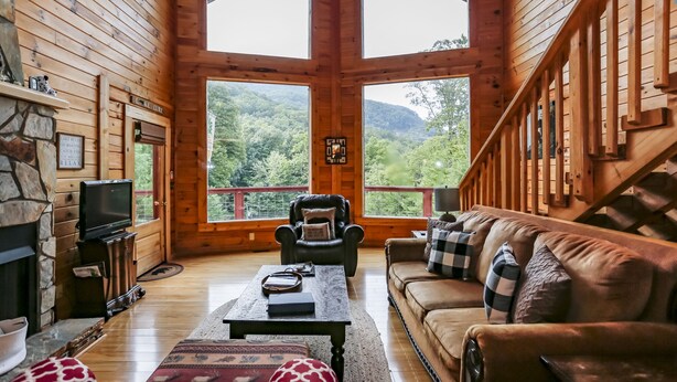 Interior d'una cabana de fusta amb una llar de foc, una televisió, un sofà i una finestra gran amb vista al bosc.
