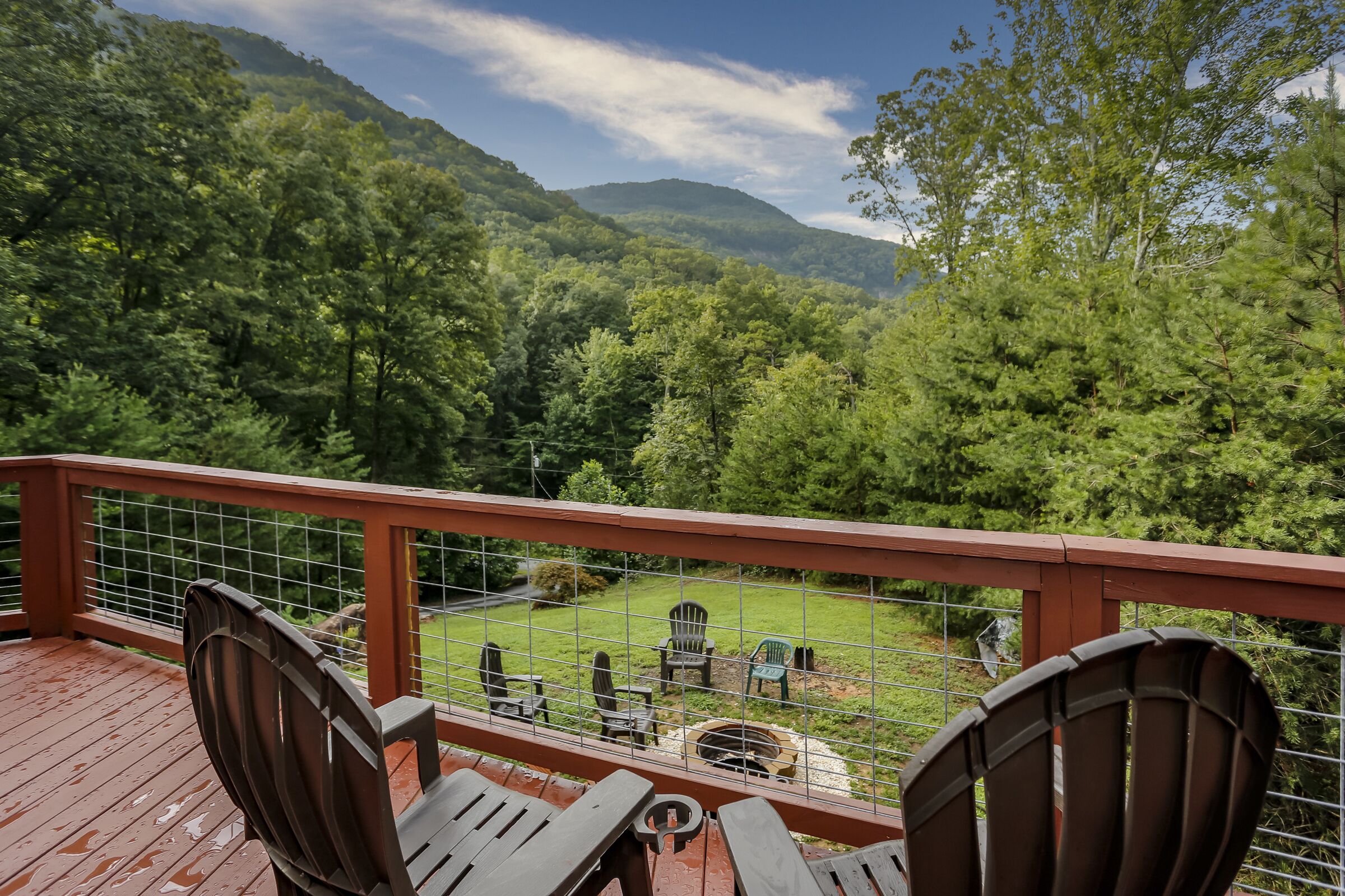 A wooden deck with Adirondack chairs overlooking a grassy yard with a fire pit and a view of a forested mountain.