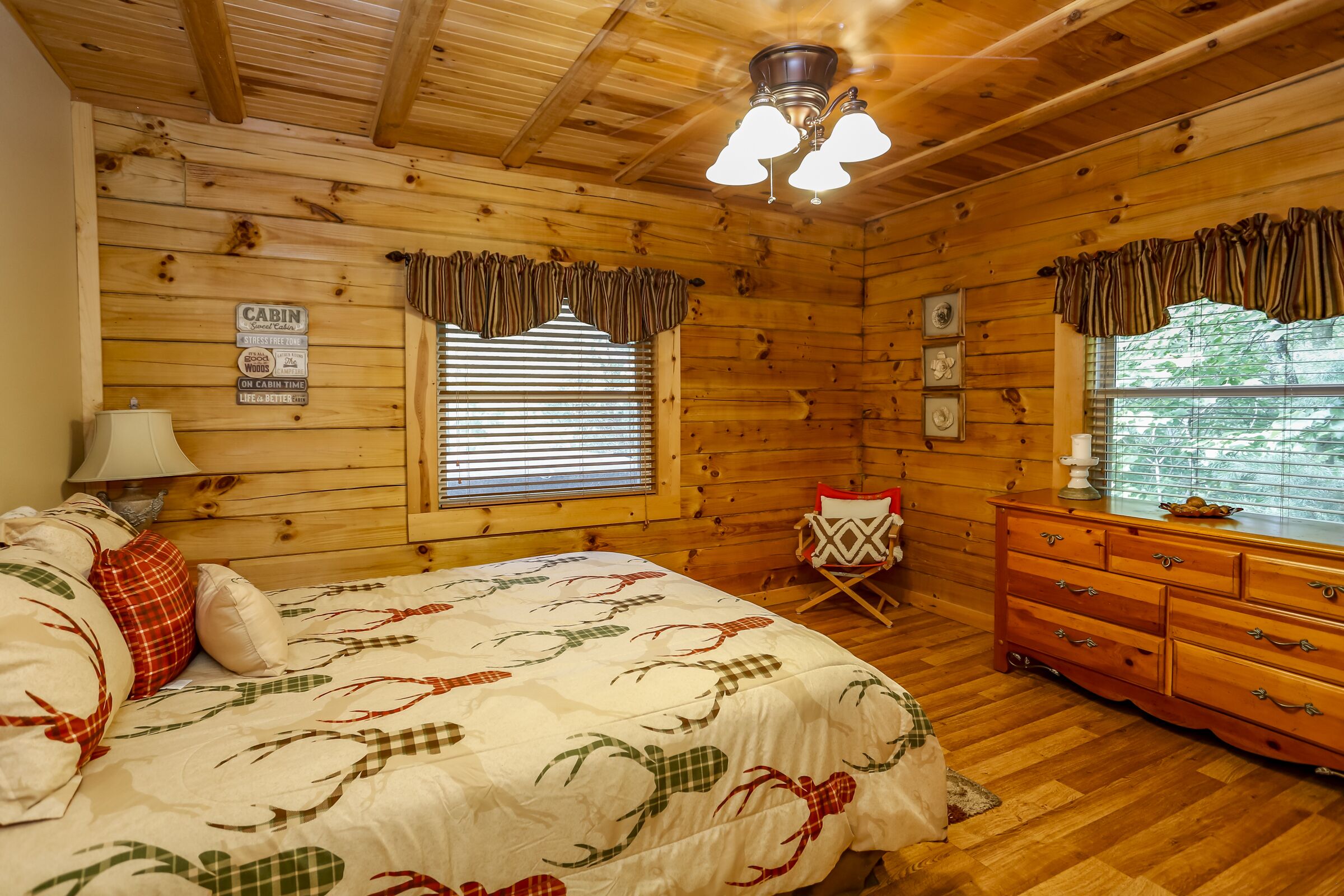 Een kamer in chaletstijl met een houten plafond, een bed met een geruit plaid, een dressoir en een raam met verstelbare zonwering.