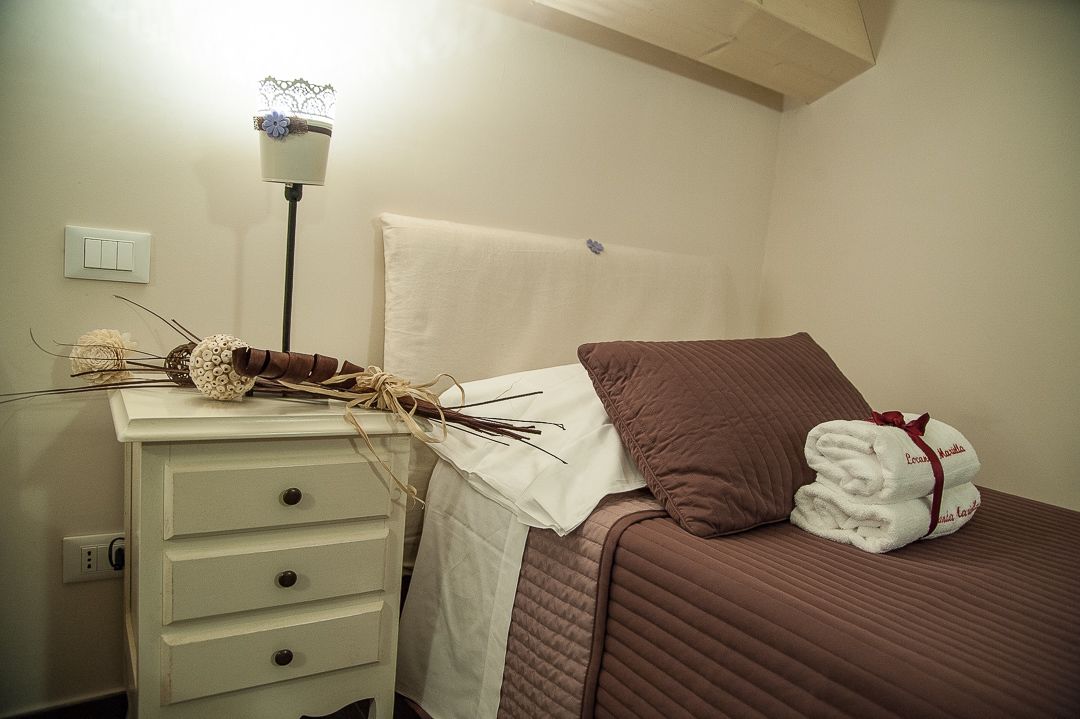 A neatly made bed with a brown pillow and a white blanket, a bedside table with a lamp and a decorative vase, and a wall-mounted light switch.