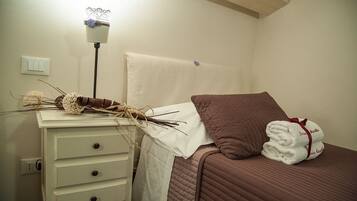 A neatly made bed with a brown pillow and a white blanket, a bedside table with a lamp and a decorative vase, and a wall-mounted light switch.