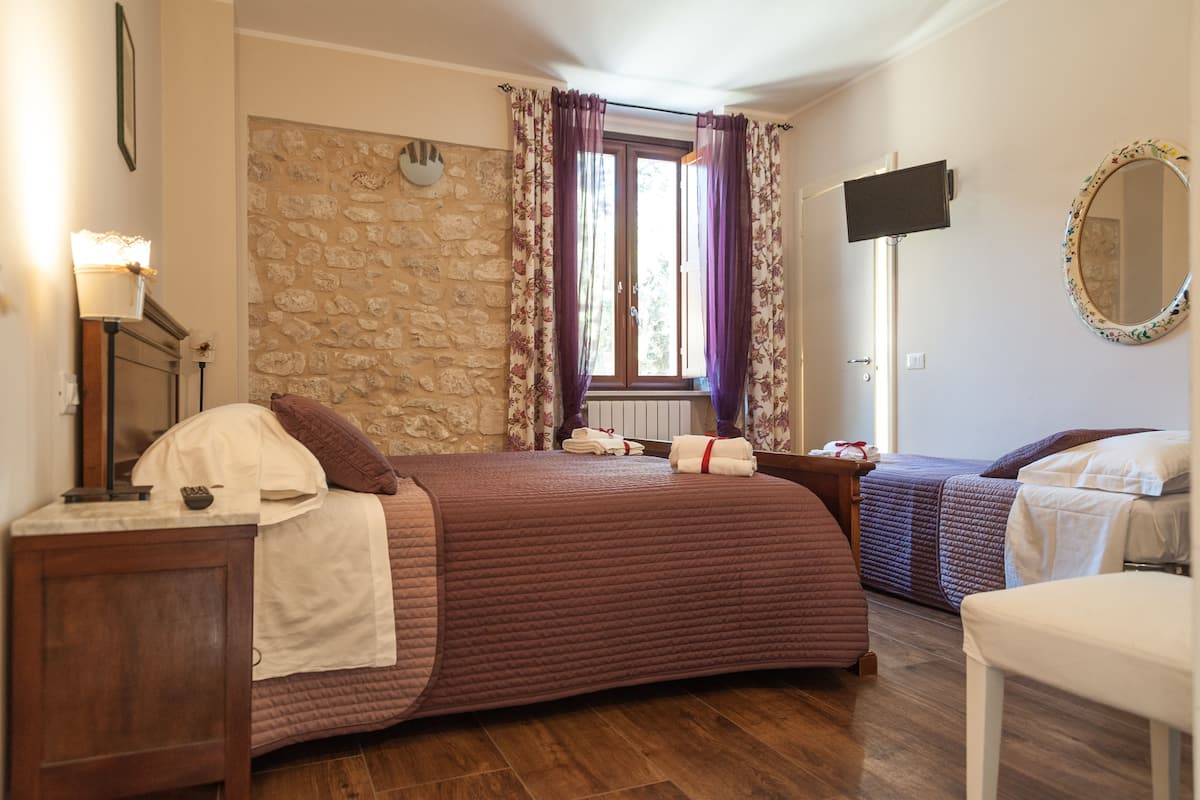 Une chambre avec un mur en pierre, un grand lit avec une tête de lit, une télévision et une fenêtre avec des rideaux.