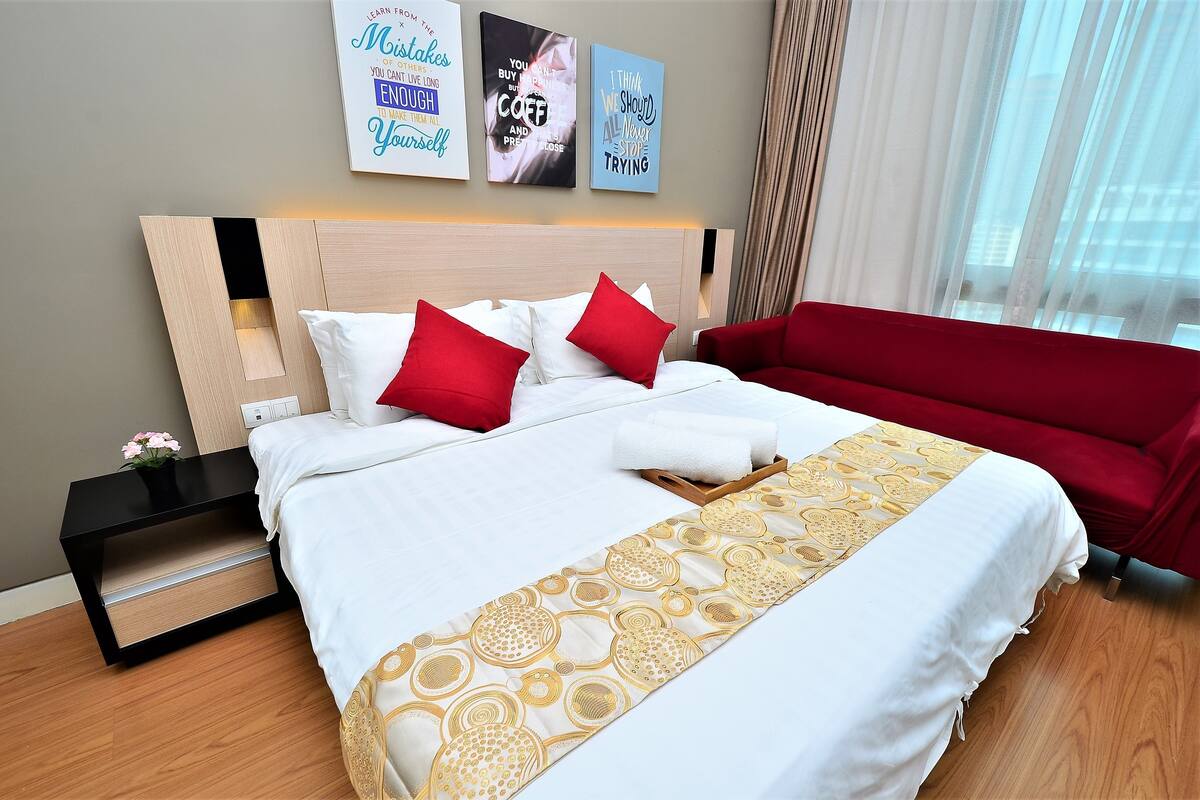 Une chambre d’hôtel avec un lit, des oreillers rouges et blancs, un canapé rouge, une table de chevet noire et trois œuvres d’art murales inspirantes.